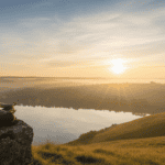 Person meditating at sunrise on a cliff, warm golden light over misty hills and a calm lake.