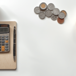 Notebook, calculator, coins, plant, and coffee on white desk.